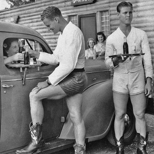 fasc1nate's tweet photo. Drive-in car hops in shorts and cowboy boots at the Log Lodge Tavern near Love Field Airport in Dallas, Texas, 1940.⁣
⁣
A woman from Oak Cliff (a Dallas neighborhood) made a complaint that she did not want to look at girls’ legs when she stopped by her local drive-in; she wanted to look at men’s legs. Drive-in owners thought that this was a great idea, and soon the idea of the scantily-clad male carhop was born.⁣
⁣
The photo above ran in newspapers around the country with the headline: “Adonis and Apollo of Roadside Bring Trade to Daring Stand.”⁣
⁣
The caption read:⁣
⁣
"First large roadside stand Friday to bow to the demand of Dallas women and feature husky young male carhops in shorts was the Log Lodge Tavern at Lemmon and Midway where four six-footers found jobs. Above, in blue shorts, white sweatshirt and cowboy boots, Joe Wilcox serves Pauline Taylor who smiles her approval of the idea. Bound for another car is James Smith, at right."