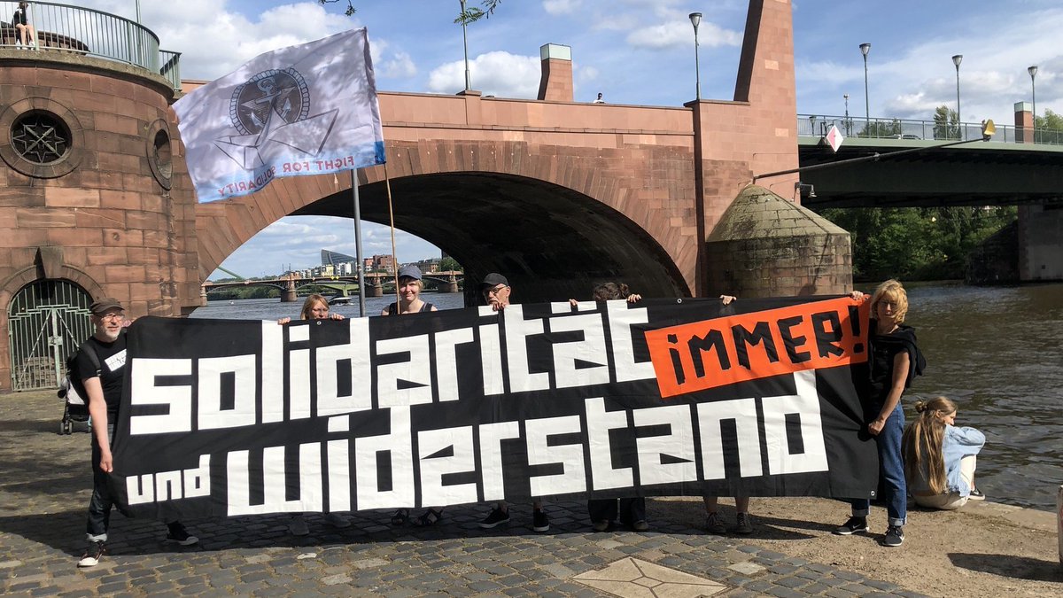 Menschen mit einem großen schwarzen Banner vor der Alten Brücke in Frankfurt.<br>Darauf steht:</p><p>SOLIDARITÄT<br>und WIDERSTAND<br>immer</p><p>1 Person hält die Iuventa-Crew-Flagge mit dem Logo und einem Papierboot und der Text: Fight for Solidarity!