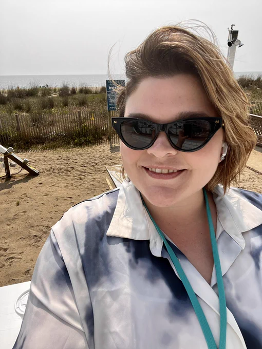 Selfie of Sam, a white woman with short blonde hair, wearing sunglasses, with the beach in the background.