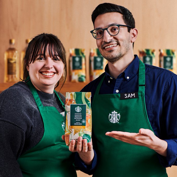 Two smiling Starbucks baristas in green aprons proudly hold a bag of Starbucks Green Apron Blend coffee.