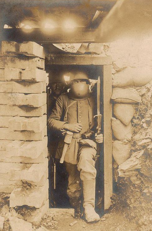 During World War I, a German soldier with a saw-tooth bayonet stands in a dugout. His helmet's brow plate, an attachment, is slid down to his neck, offering additional protection against hazards such as grenade shrapnel.