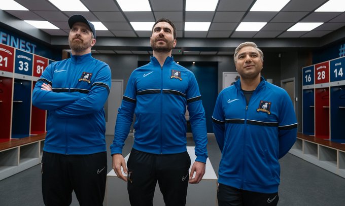 Beard, Roy Kent, and Nate stand in the locker room, wearing their new coaching uniforms, looking up at the Believe sign.