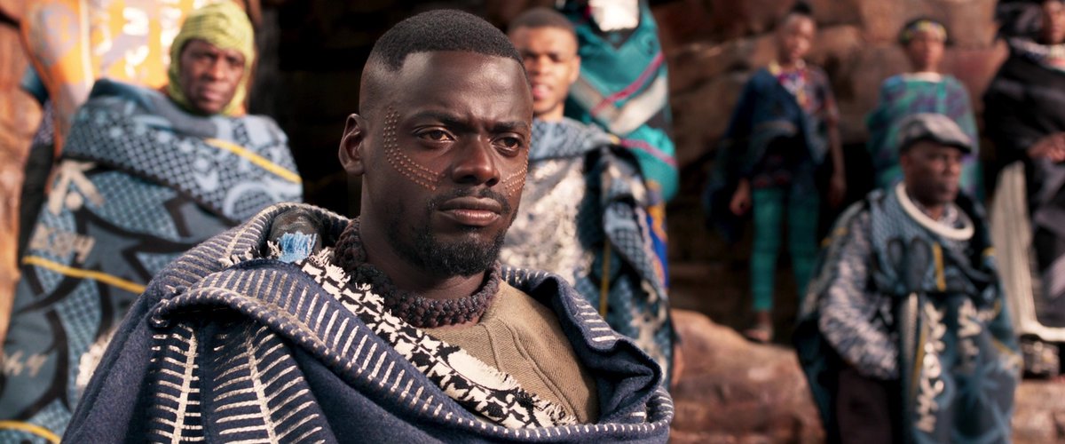 A film still from Black Panther. Daniel Kaluuya in blue clothing thoughtfully looks towards the right. There are others behind him facing the same direction as him. 
