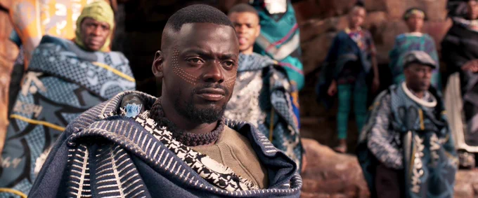A film still from Black Panther. Daniel Kaluuya in blue clothing thoughtfully looks towards the right. There are others behind him facing the same direction as him. 