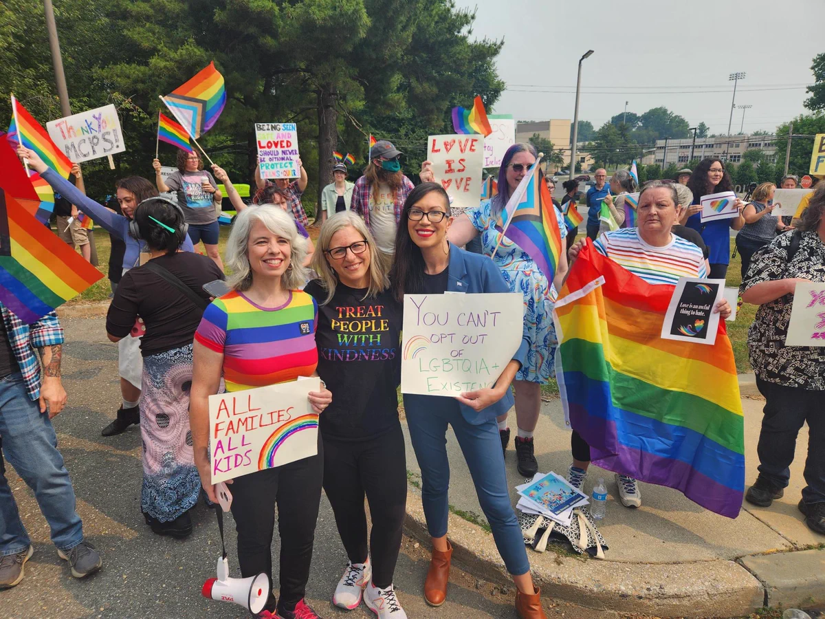 Kristin stands with Laura Stewart (MCC-PTA) and Jill Ortman-Fouse (former Board of Ed member) amongst a crowd holding rainbow signs and flags. Laura’s sign says “ALL FAMILIES, ALL KIDS.” Kristin’s sign says “You can’t opt out of LGBTQIA+ existence.”