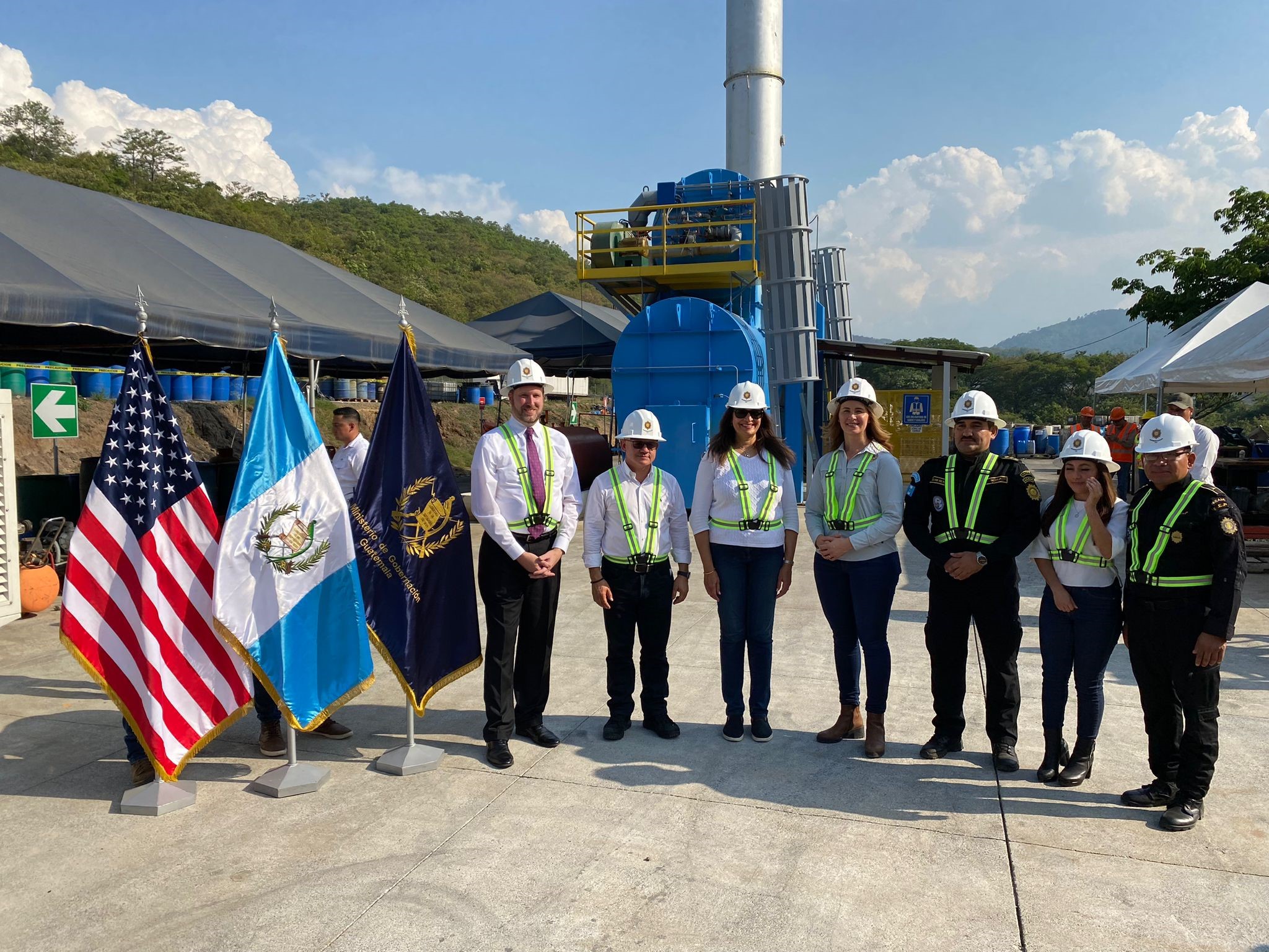 The group stands together in hard hats in front of the incinerator and next to flags.