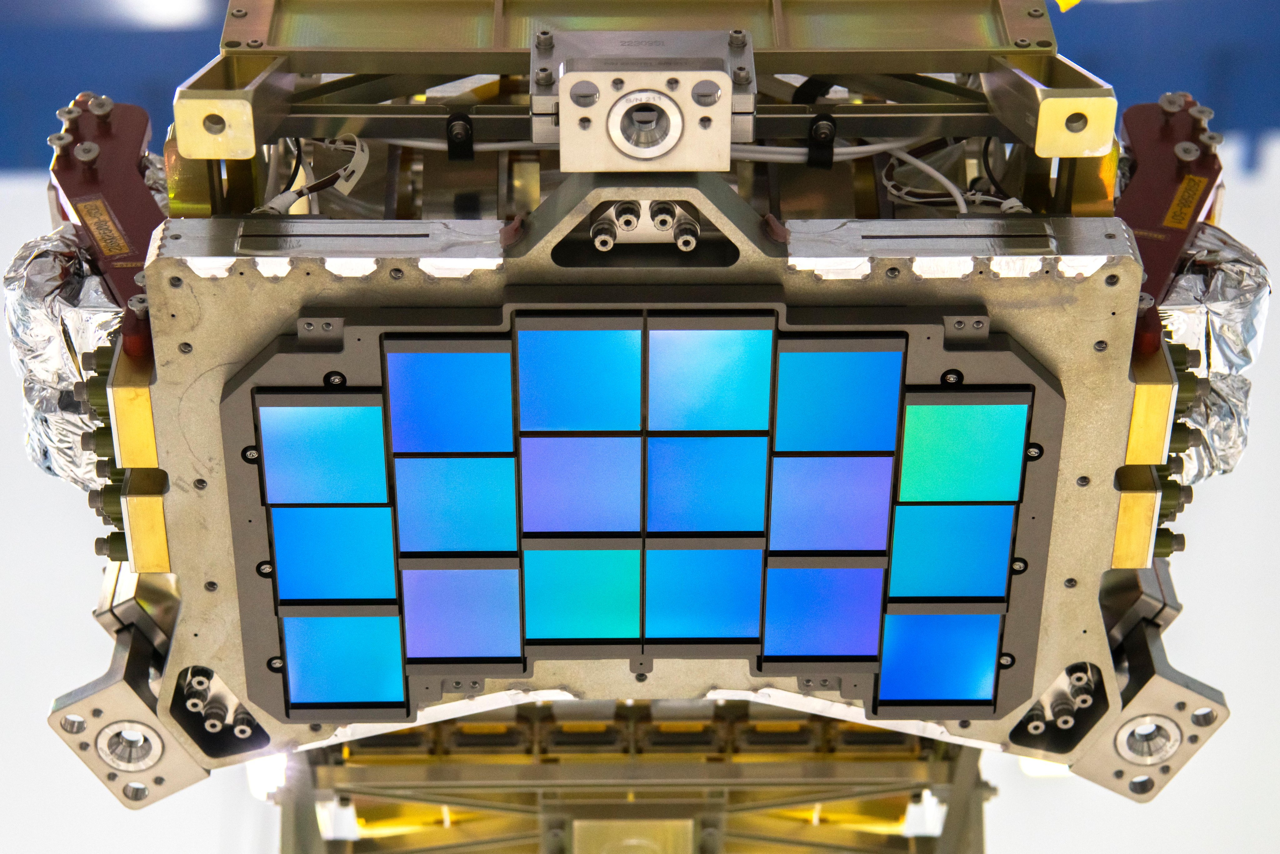 A close-up photo of an iridescent purplish-blue array of squares attached to a chunky, rectangular piece of metal hardware. The structure is about the size of a small microwave, and the array of detectors is arranged in slightly curved rows. 