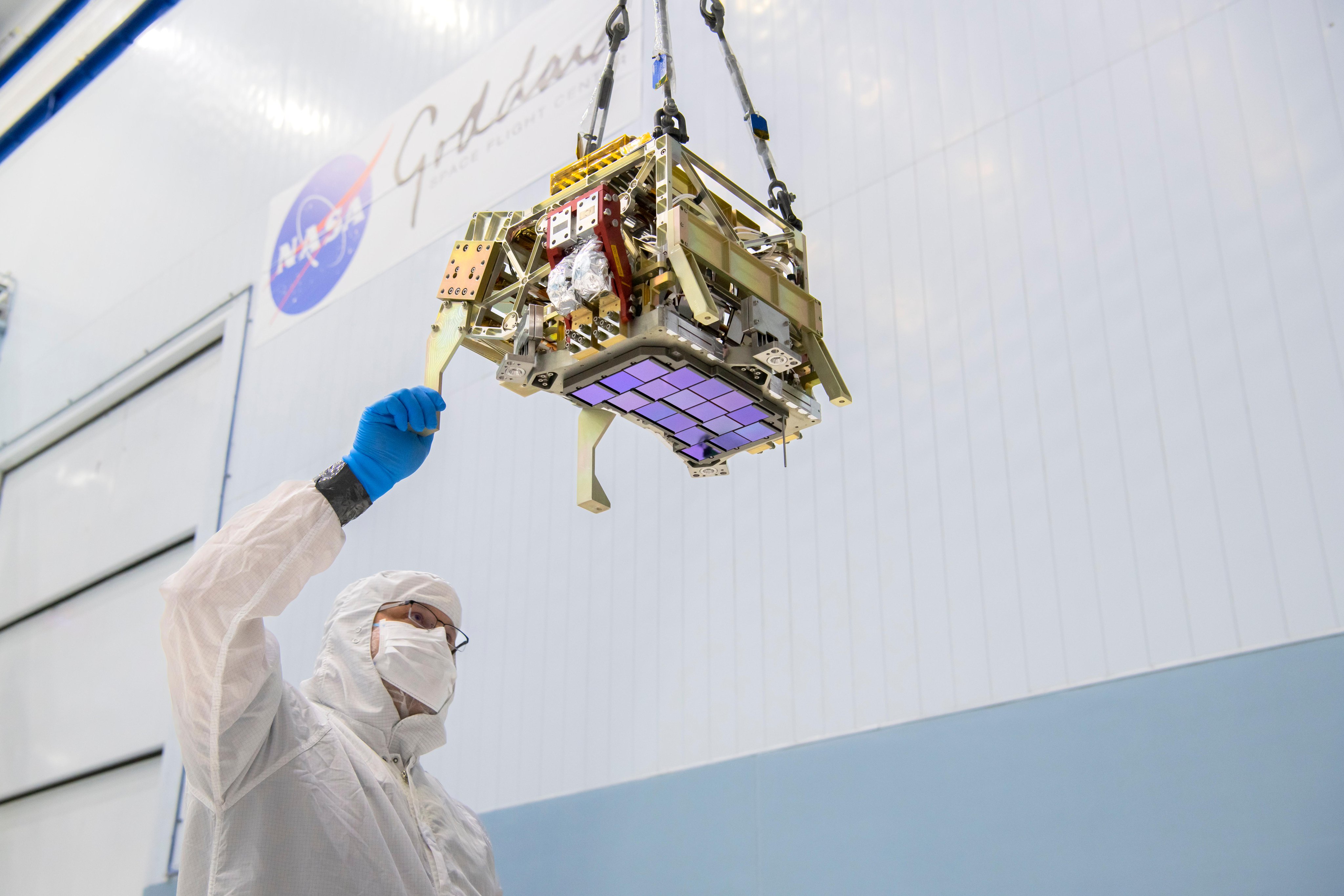 An iridescent purplish-blue array of square detectors attached to a chunky, rectangular piece of gold and silver metal hardware is seen from below. A man in a white suit and gloves holds onto an edge of the structure. The structure hangs from the ceiling of a large room with white and blue walls. It is about the size of a small microwave, and the array of detectors is arranged in slightly curved rows. Out of focus in the background, the blue and red NASA logo and text “Goddard Space Flight Center” are visible. 