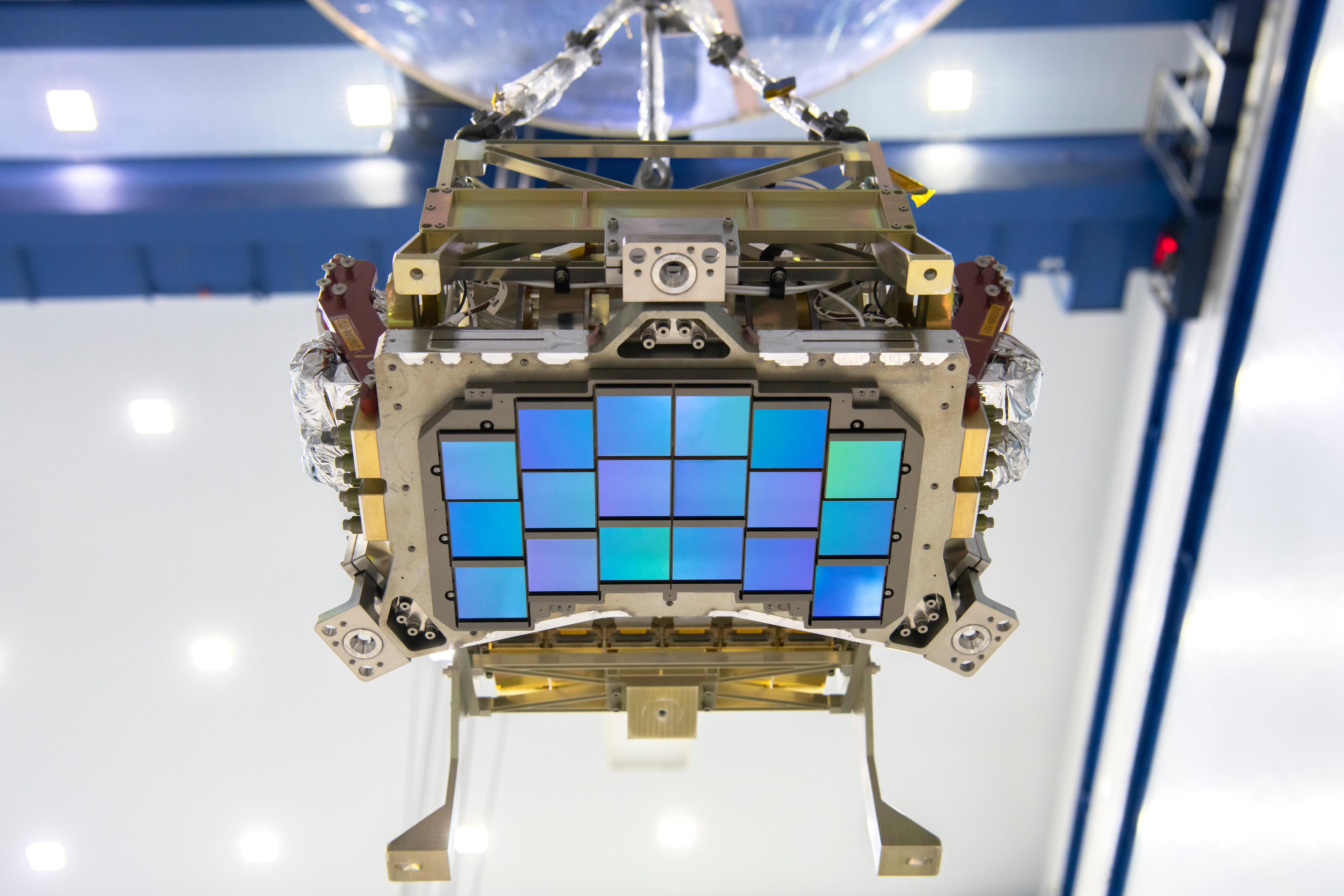 A close-up photo of an iridescent purplish-blue array of squares attached to a chunky, rectangular piece of gold and silver metal hardware, hanging from the ceiling of a large room with blue and white walls. The structure is about the size of a small microwave, and the array of detectors is arranged in slightly curved rows. 