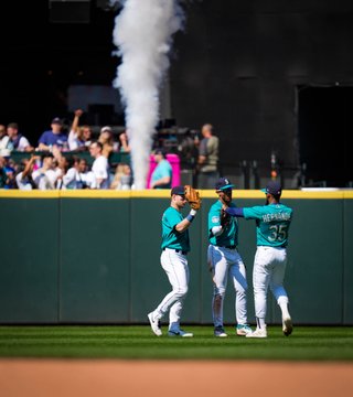 The Mariners outfield comes together in center field to celebrate the win, with fans celebrating in the T-Mobile 'Pen behind them and a column of smoke shooting up from behind the outfield wall. 