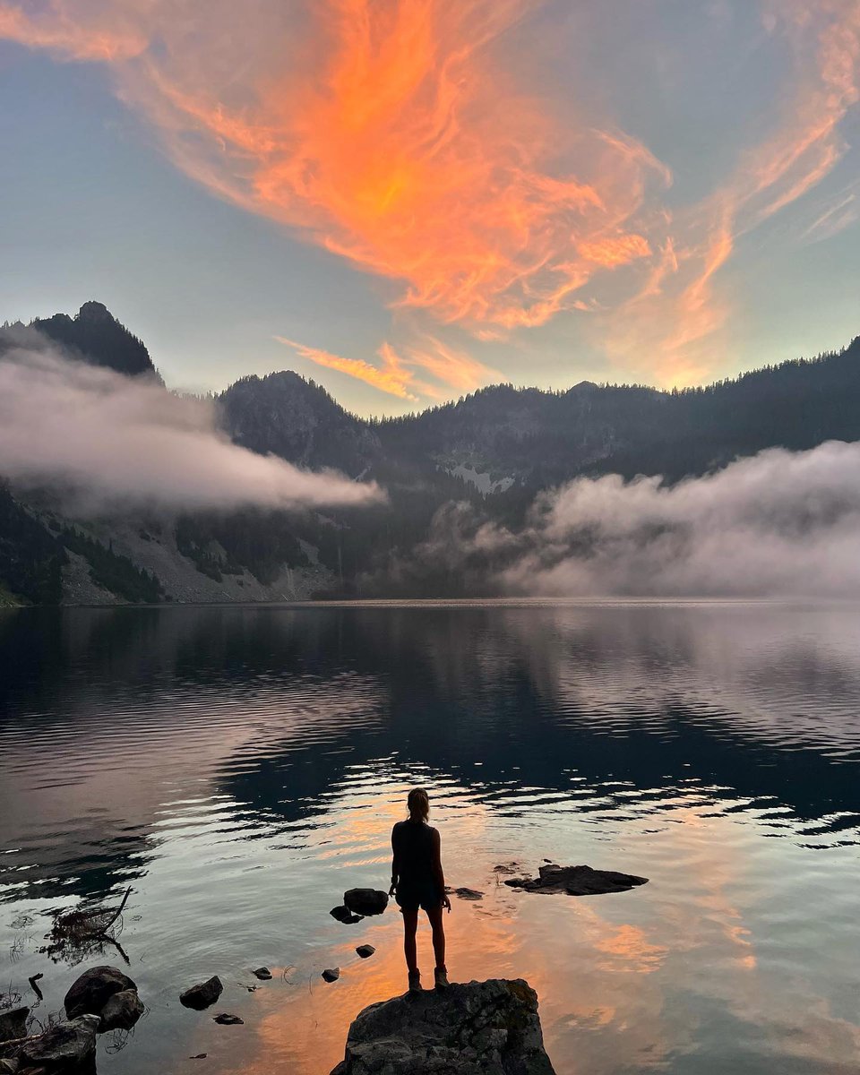 #ImageDescription: A silhouette of a hiker standing on a large rock looking ahead at an alpine lake during sunset can be seen. ( #📷 @alltrails )