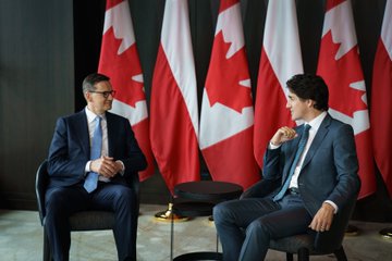 Prime Minister Justin Trudeau and Prime Minister Mateusz Morawiecki are sitting in chairs beside each other and looking at one another. Behind them, a row of Canadian flags and Polish flags is visible.