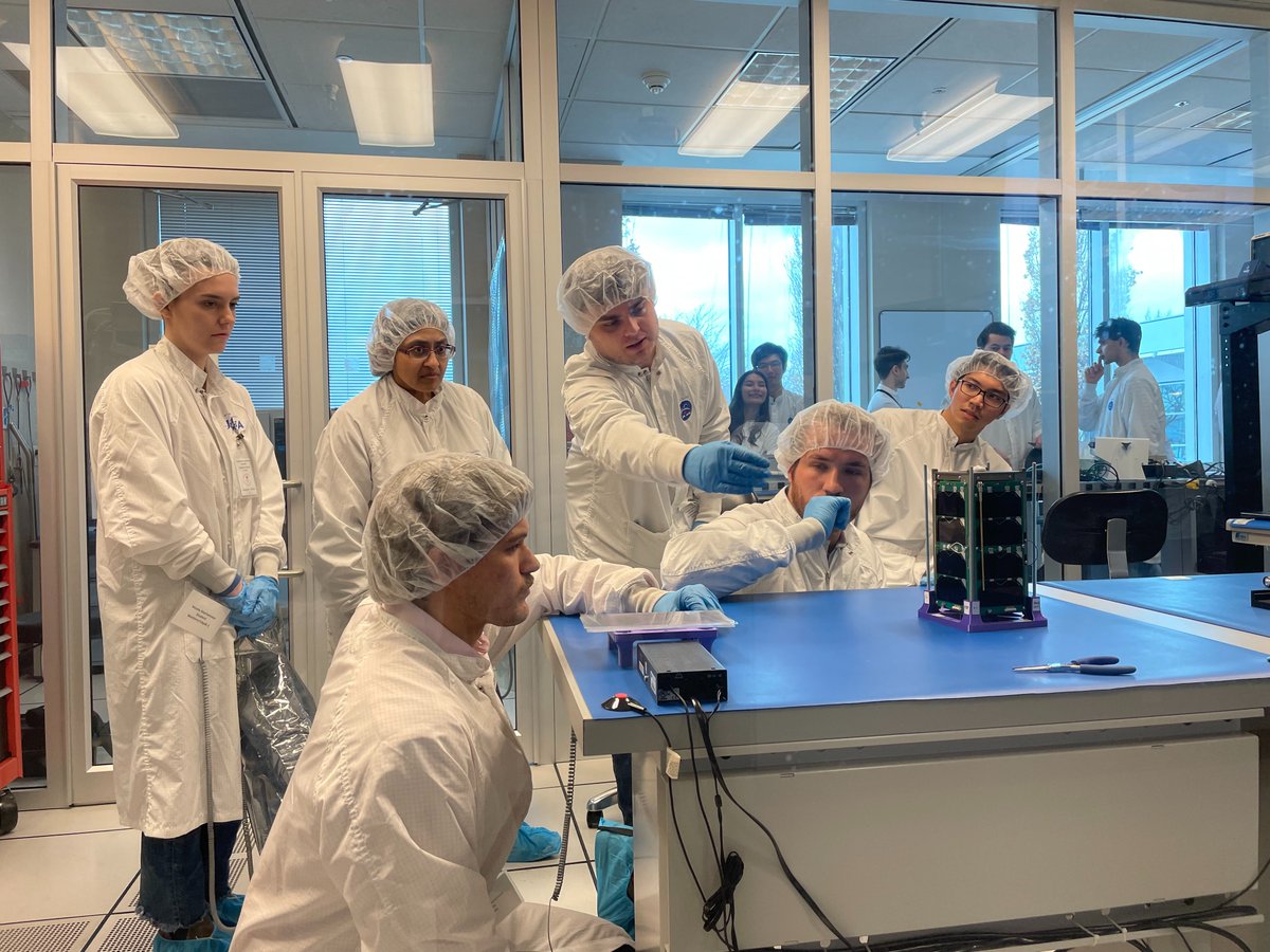 6 people in white frocks are working on a CubeSat to finalize preparation for launch. Other people are watching in the back.