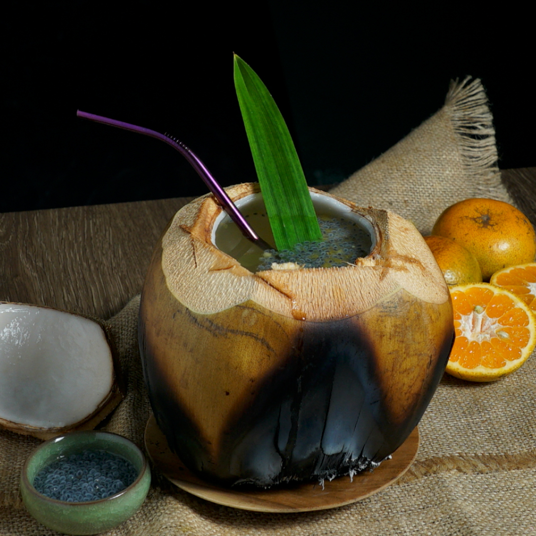 A fresh young coconut drink served in its shell with a purple straw and green pandan leaf garnish, displayed on burlap alongside oranges and a small bowl of basil seeds.