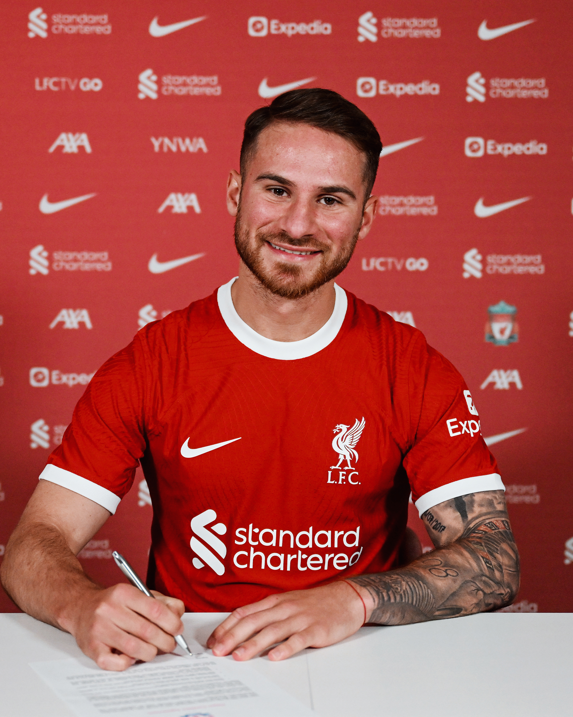 Alexis Mac Allister in the 23/24 home shirt in front of the sponsors board with a pen in his hand.