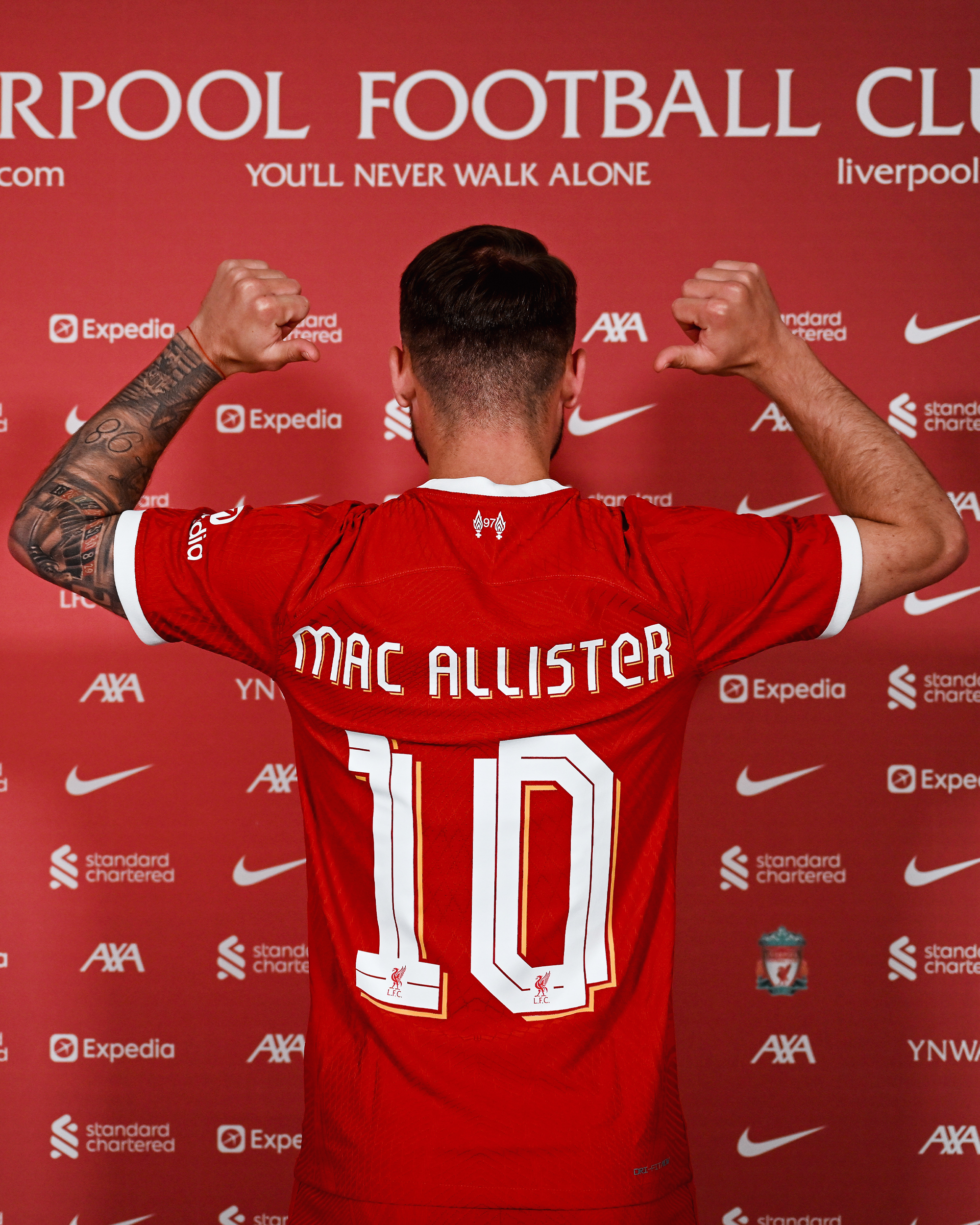 Alexis Mac Allister points to the back of his shirt in front of the sponsors board at the AXA Training Centre.