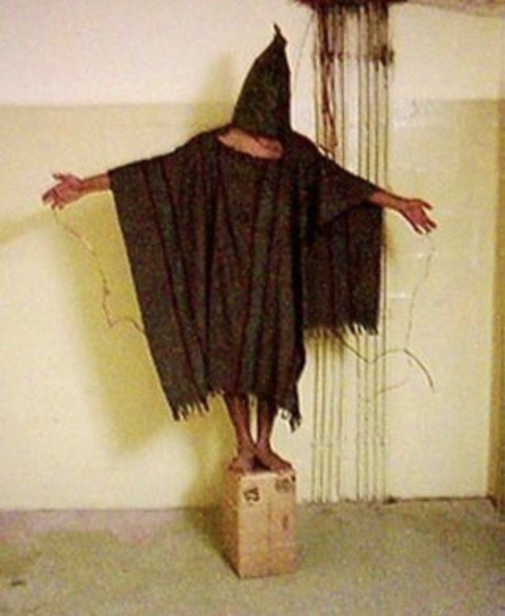 The photograph titled 'The Hooded Man' captured in 2003 depicts a distressing and unsettling scene that may resemble an eerie image often found on creepypasta or strange websites. However, it is essential to acknowledge that this photograph is an authentic representation of a