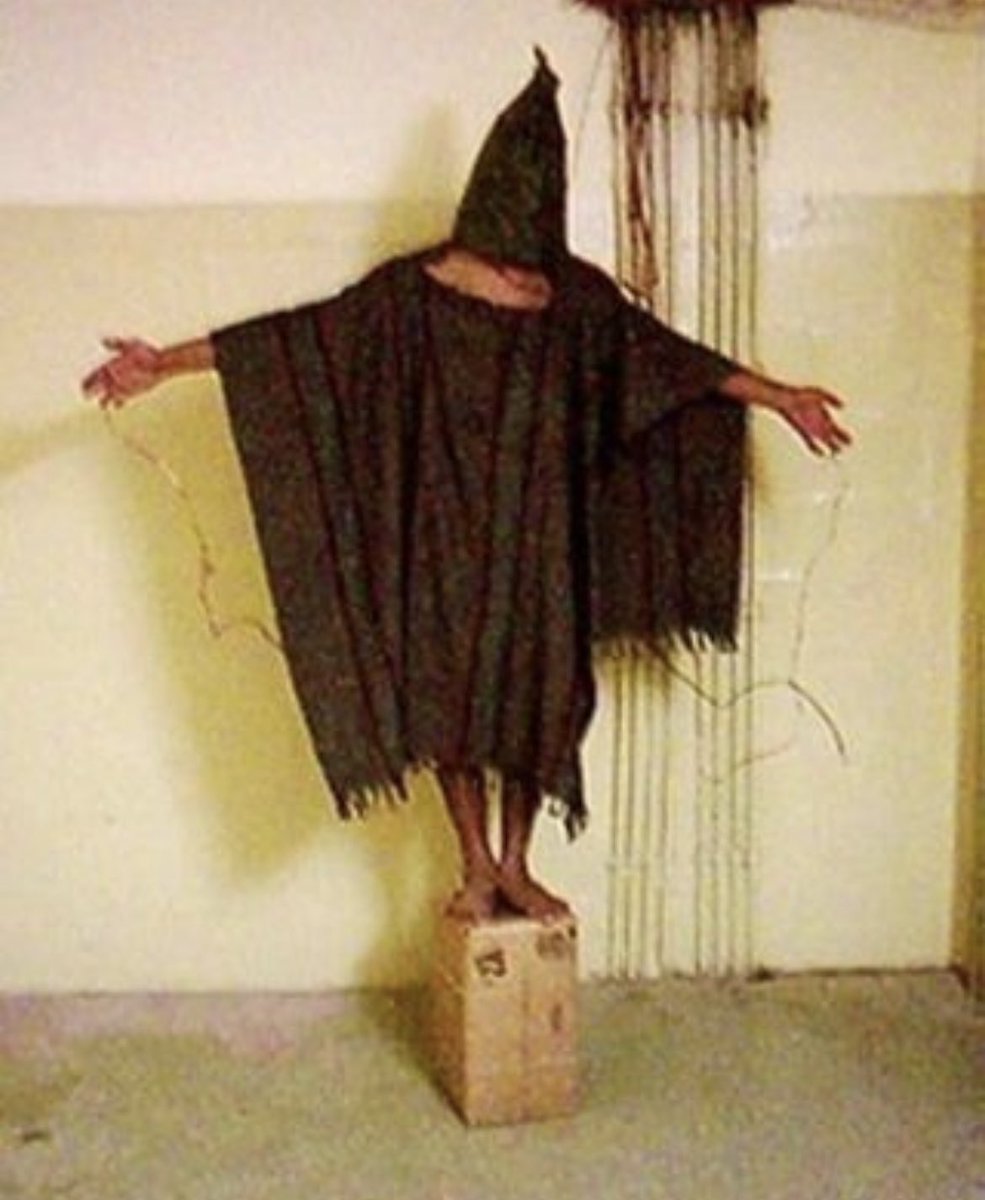 historyinmemes's tweet photo. The photograph titled "The Hooded Man" captured in 2003 depicts a distressing and unsettling scene that may resemble an eerie image often found on creepypasta or strange websites. However, it is essential to acknowledge that this photograph is an authentic representation of a real event. The image portrays an Iraqi prisoner enduring torture within the confines of Abu Ghraib prison. It is worth noting that this particular picture is relatively less severe compared to a series of photos illustrating US soldiers engaging in acts of torture against Iraqi detainees.

Originally employed by Saddam Hussein as a symbol of terror, Abu Ghraib prison later became a facility utilized by the United States to detain suspected insurgents. Staff Sergeant Ivan Frederick, responsible for capturing this photograph, documented thousands of images during his time at the prison.

The imprisoned individual shown in the photograph is subjected to both psychological and physical torment. Stripped of his sight, clothing, and dignity, he appears to have electric wires affixed to his fingers, hinting at the possibility of electrocution. Additionally, he is compelled to stand on a cardboard box, assuming a posture reminiscent of a crucifixion.

The prisoner's name is Ali al-Qaisi. He was wrongfully arrested on suspicion of being an insurgent after he voiced his complaint against American soldiers who had discarded refuse on a football field in his town. Although Qaisi was known to hold sympathies for the insurgents, US authorities apprehended him for further questioning. Upon his arrival at the prison, Qaisi described the deplorable treatment he and other detainees endured, stating, "As soon as one enters the prison they hang them on the door of the metal cell, then they electrocute the person, and urinate on them".

It is important to recognize that Ivan Frederick, the photographer responsible for capturing this image, has since been identified as a war criminal. Following investigations, a total of 11 staff members from the prison were arrested. Frederick himself received an eight-year prison sentence, although he served only four years of his sentence.