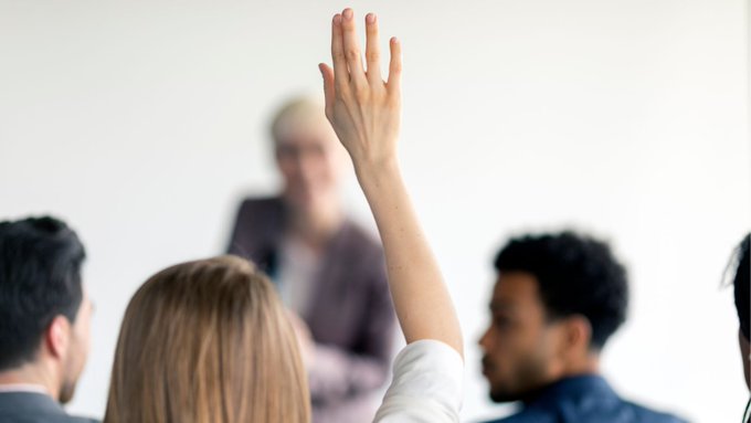A photo of someone raising their hand to ask a question. Other people are blurred in the background.