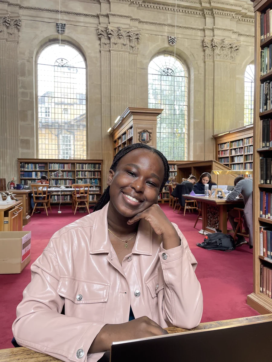 Rawletta Barrow is sitting at a desk in a library with a high ceiling and stone walls. There are large arch shaped windows behind her and a red carpet. She is leaning her chin on the back of one hand and smiling at the camera. She is wearing a pale pink leather jacket.