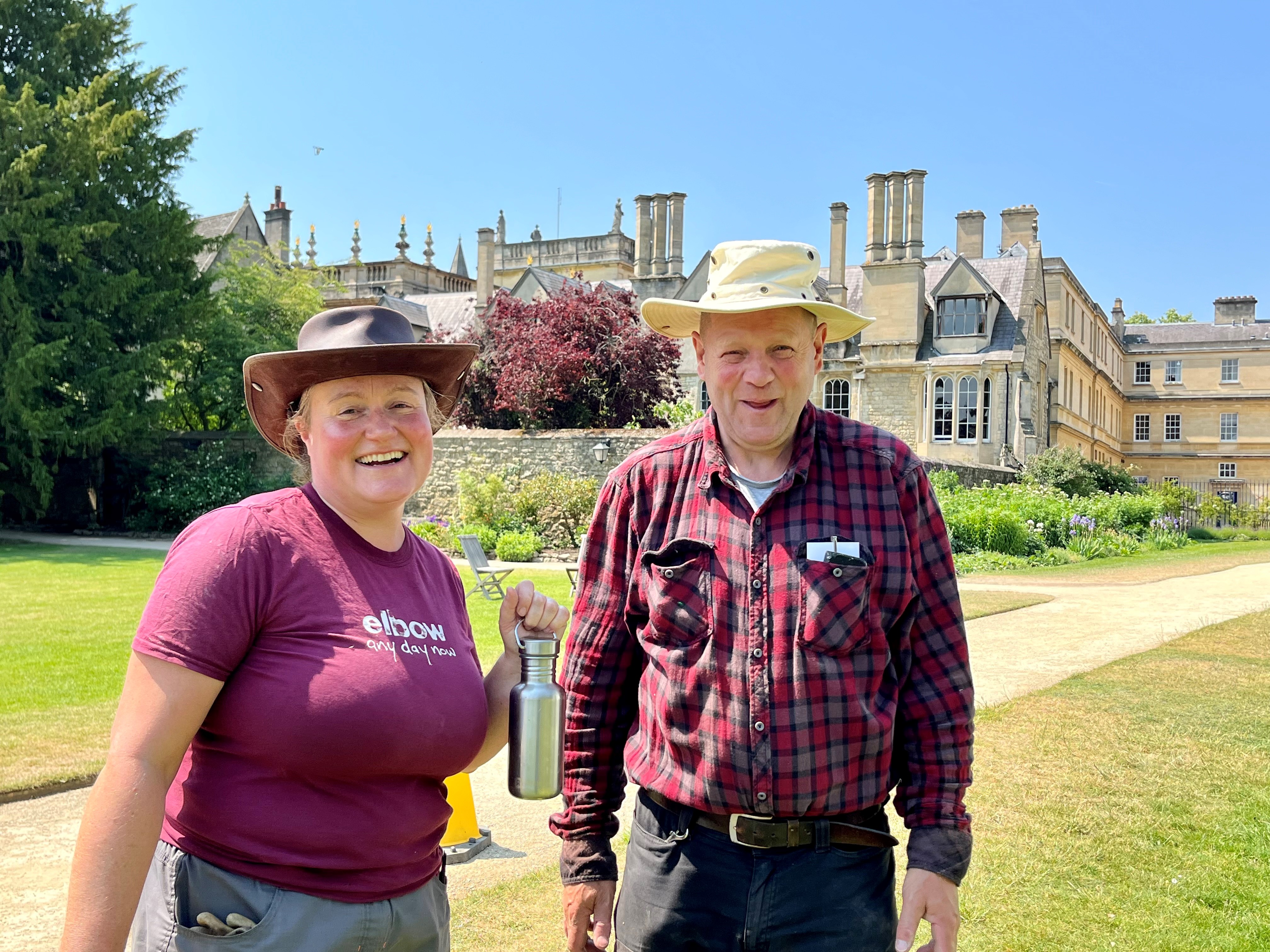 College gardeners Kate and Bob stand in the trinity lawns wearing sun hats and smiling. Kate is holding a bottle of water.