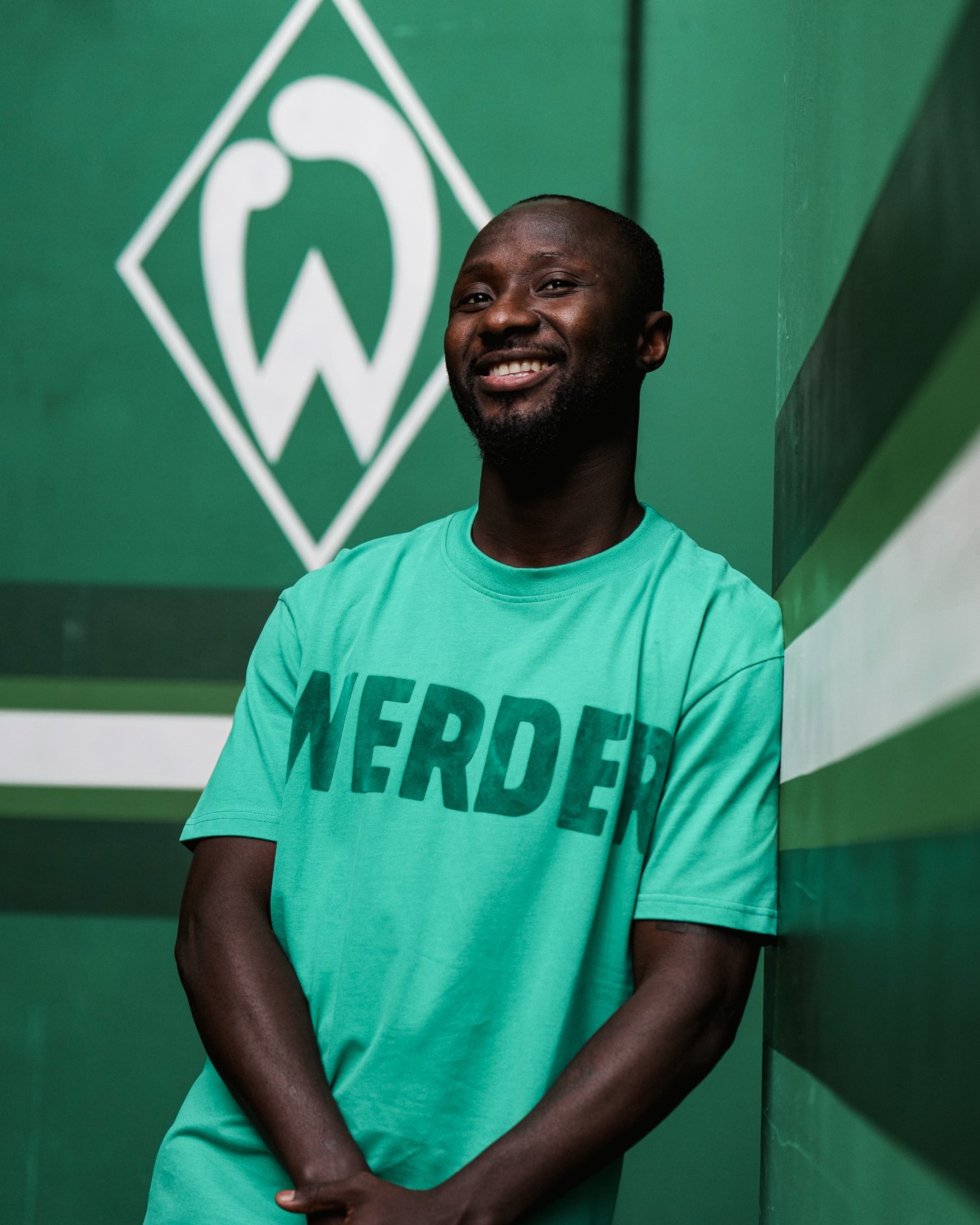 Naby Keita vor einer Werder-Raute an der Wand lehnend. 