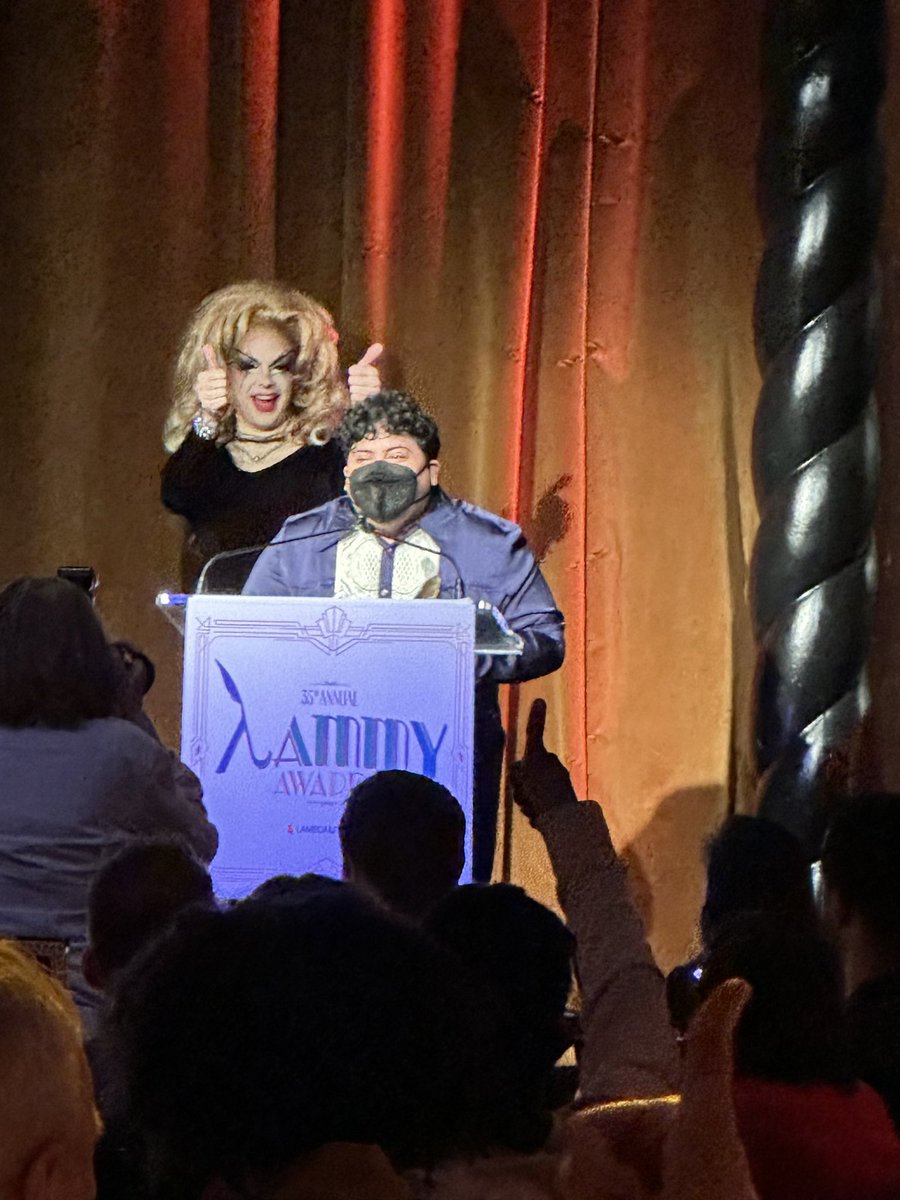 Me standing at a podium with a drag queen behind me giving a double thumbs up