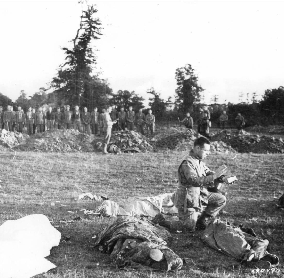 While a burial party of German POWs awaits to complete their assignment, Paratrooper and Regimental Chaplain with the 101st Airborne (501st PIR), Father Francis Sampson, gives last rites to paratroopers killed in the Battle of Carentan in June of 1944. 🪂