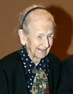 Mary Zelia Pease Philippides, very late in life; a smiling older white woman with white hair, wearing a navy hairband and a polka-dot blouse with a dark blazer