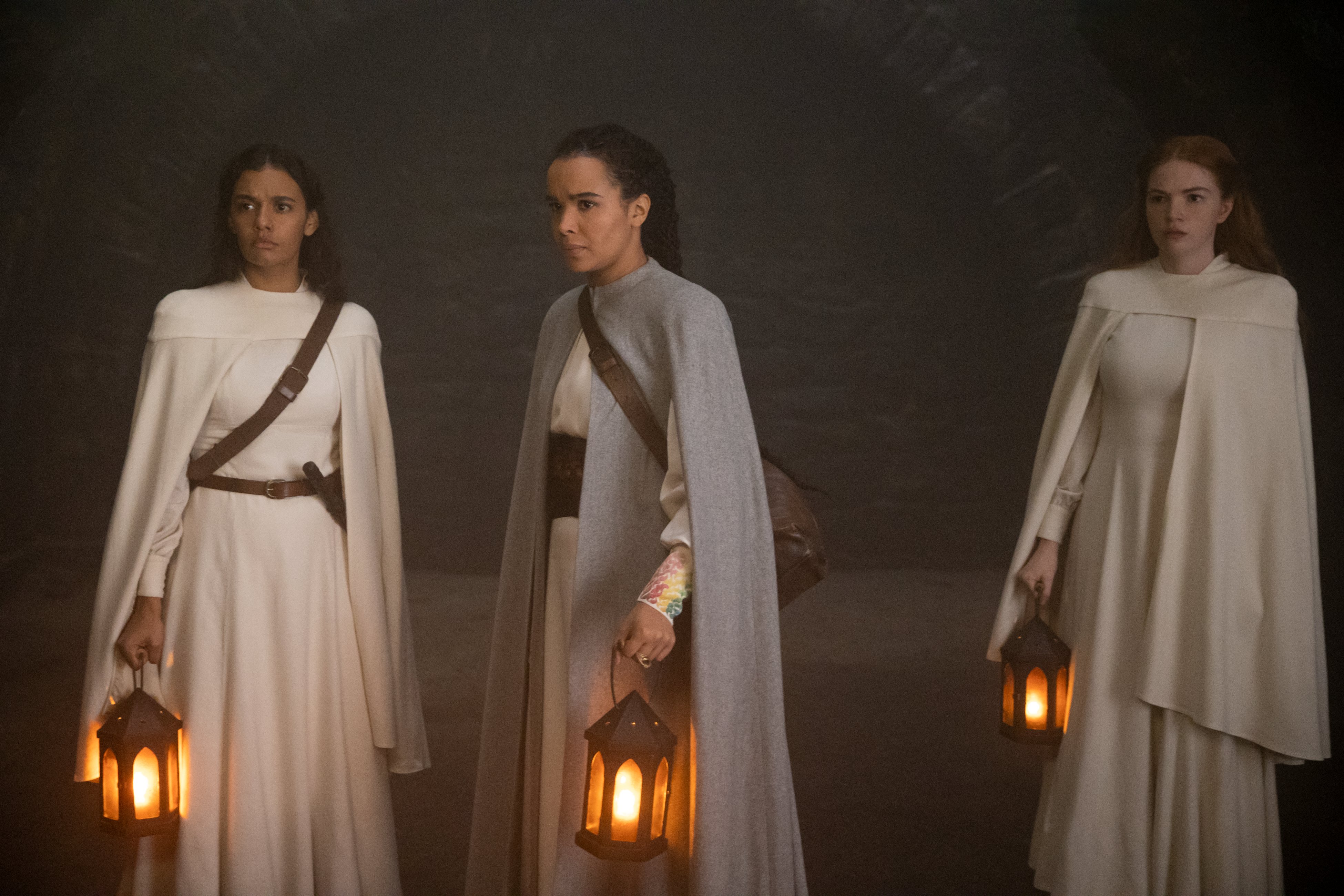 Full shot of Egwene, Nynaeve, and Elayne standing in a dark cave, wearing their outfits from The White Tower. Each carries a glowing lantern. Egwene and Nynaeve wear leather over-shoulder weapon holsters.