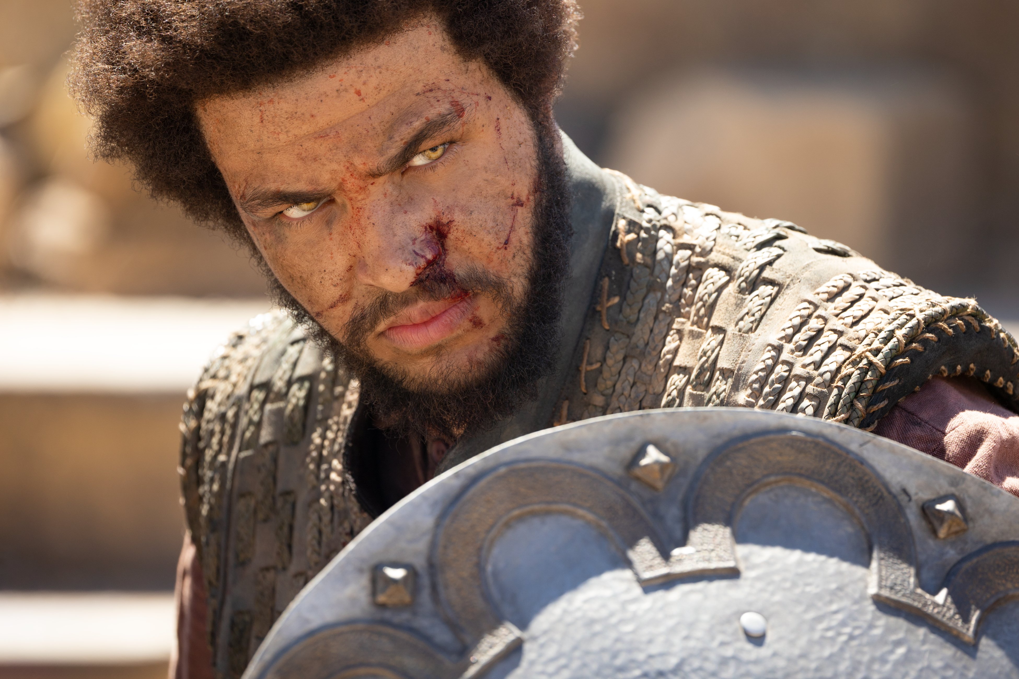 Close-up shot of Perrin looking intensely at something off camera, his face bloodied from battle. He holds up a shield to his shoulder. His eyes glow golden yellow.