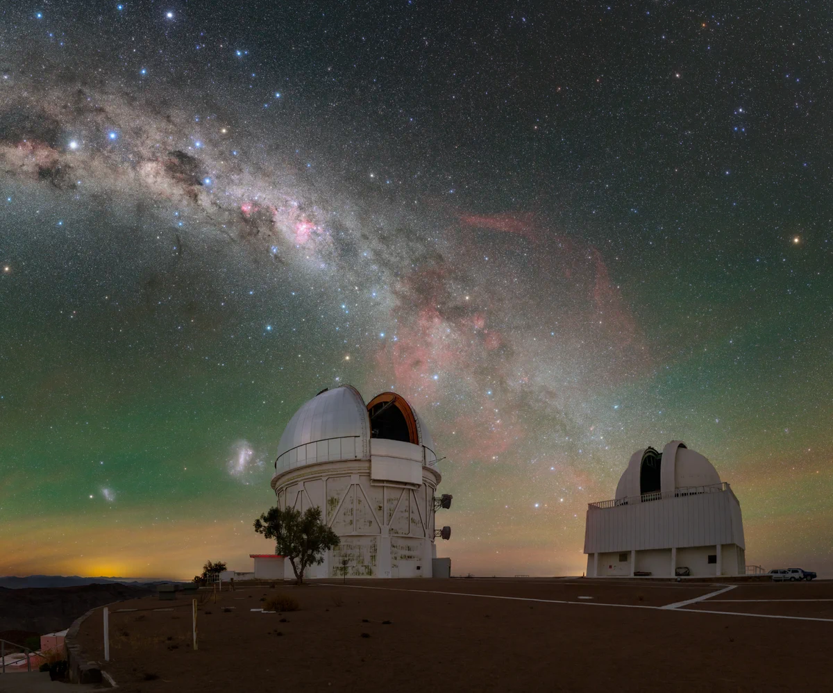Speckled with multi-colored stars, our home galaxy in this image stretches down into the city-lit horizon, just grazing past the Víctor M. Blanco 4-meter Telescope (left) and SMARTS 1.5-meter Telescope (right). The Small and Large Magellanic Clouds that accompany our home galaxy are seen to the left of the Blanco telescope. In between the telescopes blooms a deep shade of red that permeates through the tendrils of the Milky Way. This is the Gum Nebula, an emission nebula that blazes with the light of gas ionized by its host stars.