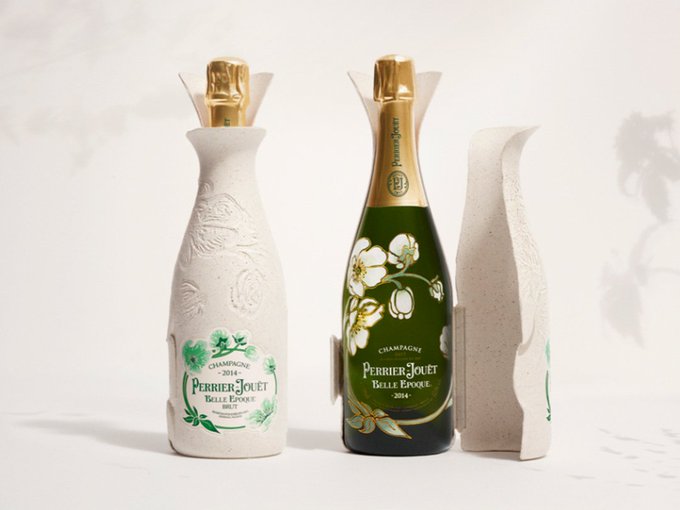 A photograph of two glass bottles of Perrier-Jouët Belle Epoque. The bottle on the left is wrapped in the Perrier-Jouët Belle Epoque Cocoon, a gift wrap made of paper pulp and vine shoots. It is white and embossed with a flower sign. The bottle to the right is also wrapped in the Cocoon, but it has been opened to reveal the green glass bottle inside, with the lid and neck wrapped in a gold foil. A similar floral design has been printed onto the bottle, now coloured with green stems and white flowers. 