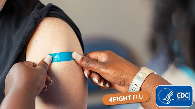 A health care professional with manicured hands places a bright blue bandage on a patient’s upper arm after they received a flu vaccine. In the bottom right-hand corner, text overlay on an orange bandage says “FightFlu” and is next to the CDC/HHS logo.   