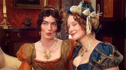 Caroline Bingley whispers to Louisa Hurst. Caroline wears an over trimmed orange empire gown and Louisa wears an over trimmed blue gown with a turban. From Pride and Prejudice (1995).