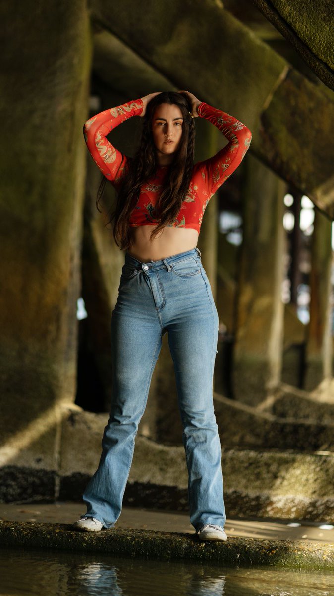 CzoborN's tweet photo. "Beneath the industrial shadow of a concrete pillar pier, a Latina muse commands attention. Clad in a vibrant red top that ignites passion, and adorned with blue jeans that embody resilience, she embodies an unwavering attitude that refuses to be subdued." https://t.co/UODVlB8gnl