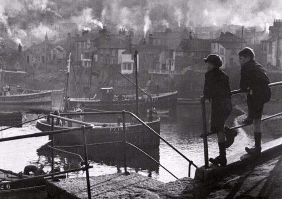 This nostalgic photo taken by Geraldine Underell in the 1940’s evokes memories and thoughts of bygone Mousehole, Cornwall Blighty days. It shows village boys Dick Cary and Donald Waters watching local fishermen hard at work in the harbor or down the ‘por’ as it was known locally.