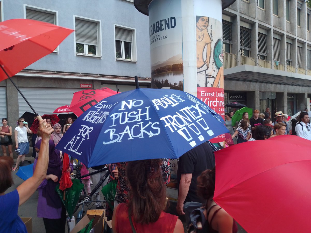 Menschen mit teilweise beschrifteten Regenschirmen vor dem Bürgeramt in Mainz. Im Zentrum des Bildes ein blauer Regenschirm. In den drei sichtbaren Feldern steht in weißer Schrift:<br>EQUAL RIGHTS FOR ALL<br>NO PUSH BACKS<br>ABOLISH FRONTEX !!!