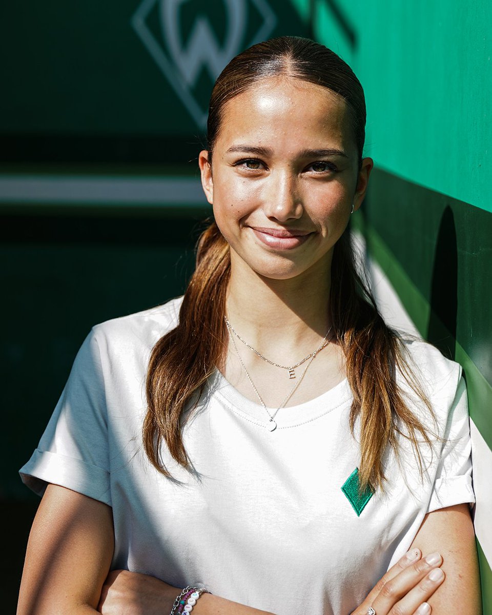 Amira Dahl trägt ein weißes Tshirt und lehnt mit verschränkten Armen an einer grünen Wand.