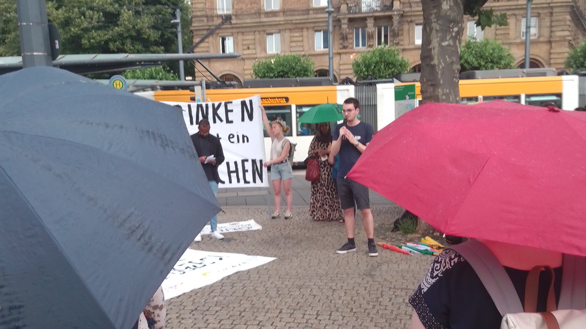 Bahnhofsvorplatz in Mainz. Blick durch zwei Regenschirme: Stefen von Resqship spricht in ein Mikrofon. Im Hintergrund halten Menschen ein Banner ERTRINKEN lassen ist ein VERBRECHEN