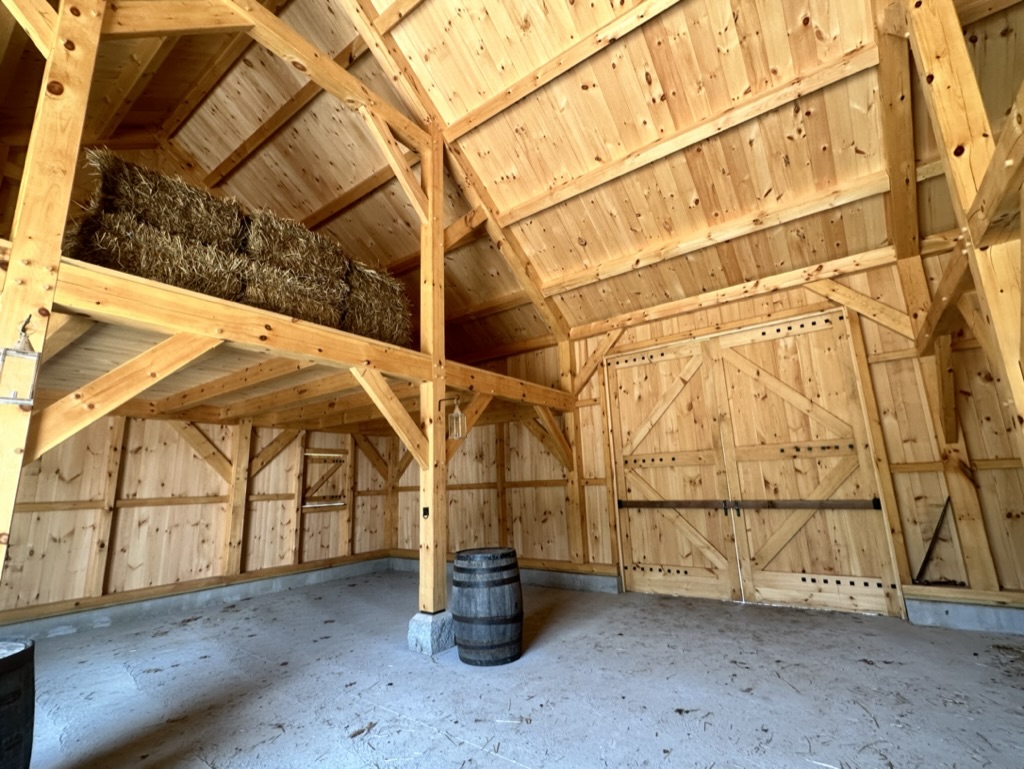 We love this view inside our new threshing barn! This was built as an early 19th-century reproduction barn, using post and beam construction. Isn't it stunning?!

#AltText: A wooden barn with a hay loft, concrete floor, and timber frame.