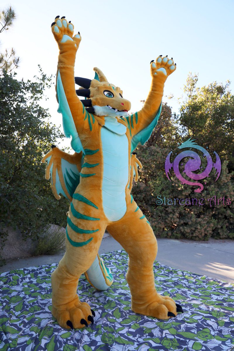 A dragon fursuiter raising his arms into the sky. The dragon is a rich orangey yellow color with lots of blended green accents and stripes.