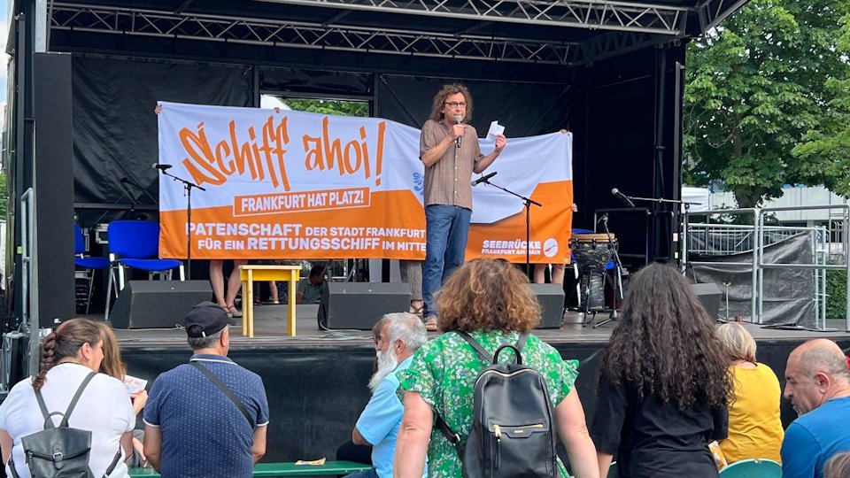 Auf einer Bühne an der Bockenheimer Warte halten Personen ein orange-weißes Transpi hoch: SCHIFF AHOI - Frankfurt hat Platz!<br>Davor spricht ein Mensch vor Publikum in ein Mikrofon.