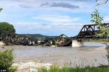 The image is taken from the river banks downstream of the wreckage. Grass and shrubs in the foreground, then muddy, turbulent river water. The bridge and train run across the middle, and in the center, you can see yellow-white froth, where sulfur seems to be leaking into the water. In the background, forested hills under a partly cloudy sky. 