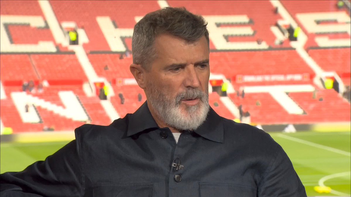 A grey-bearded man in a dark collared shirt speaking in what appears to be a stadium broadcast, with red stadium seating behind him displaying partial text 'CHE... SE... NI... T...'.