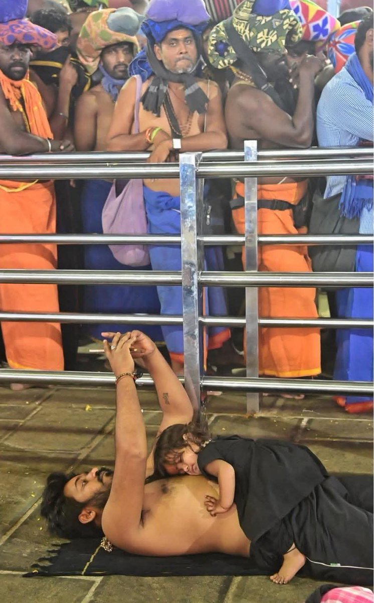 पिता की गोद में…🧡
भीड़भाड़ वाले सबरीमाला नादपंडाल के अंदर,
मंत्रोच्चार, पसीने और थकावट के बीच,
नन्ही मलिकप्पुरम अपने पिता की छाती पर शांति से सो रही है।
न बिस्तर, न आराम, फिर भी दुनिया की सबसे सुरक्षित जगह, पिता ठंडे फर्श पर लेटे हैं, दर्शन के बाद थके हुए हैं, लेकिन हिलते नहीं।