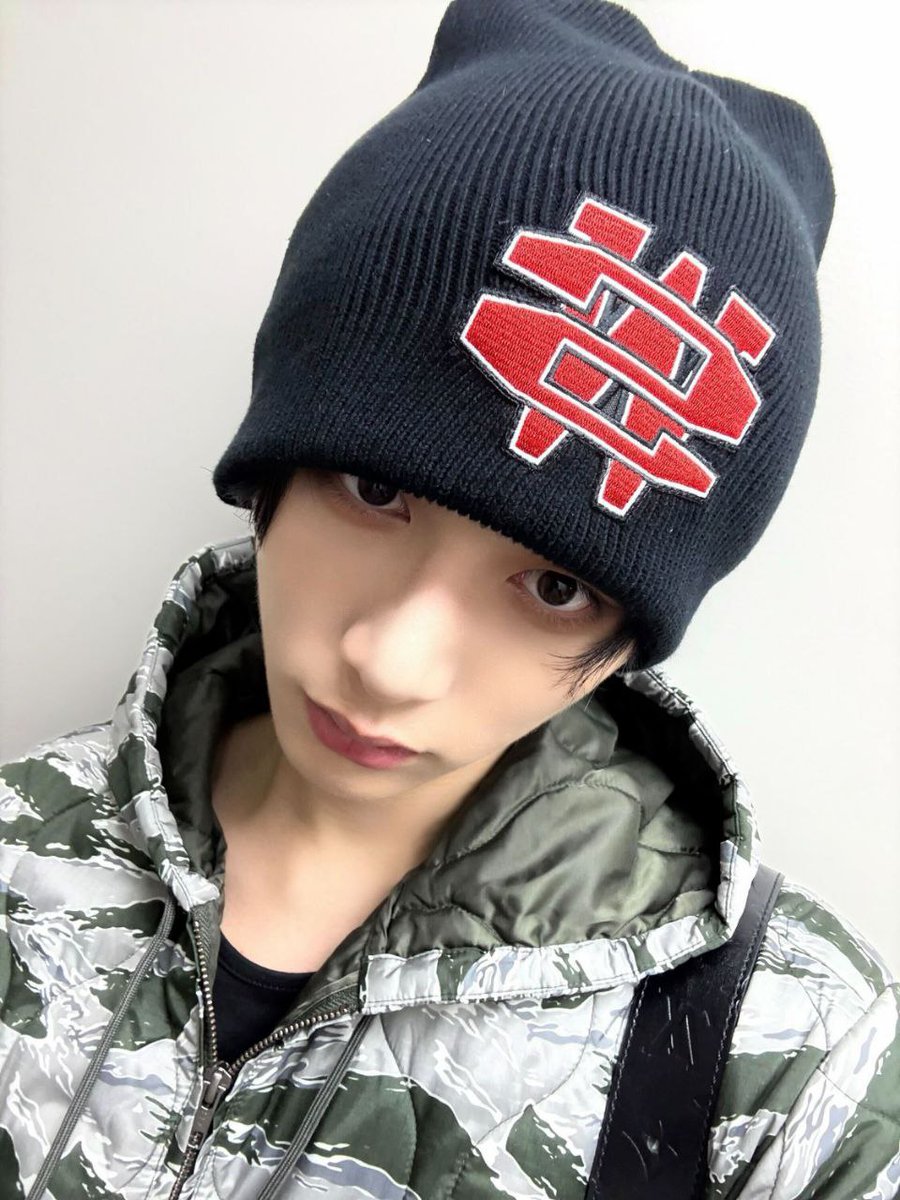 His eyes speak first beneath the stitched emblem. Patterned outerwear and a casual gesture define a modern edge, where youth shows itself through focus, restraint, and a clear sense of authority.