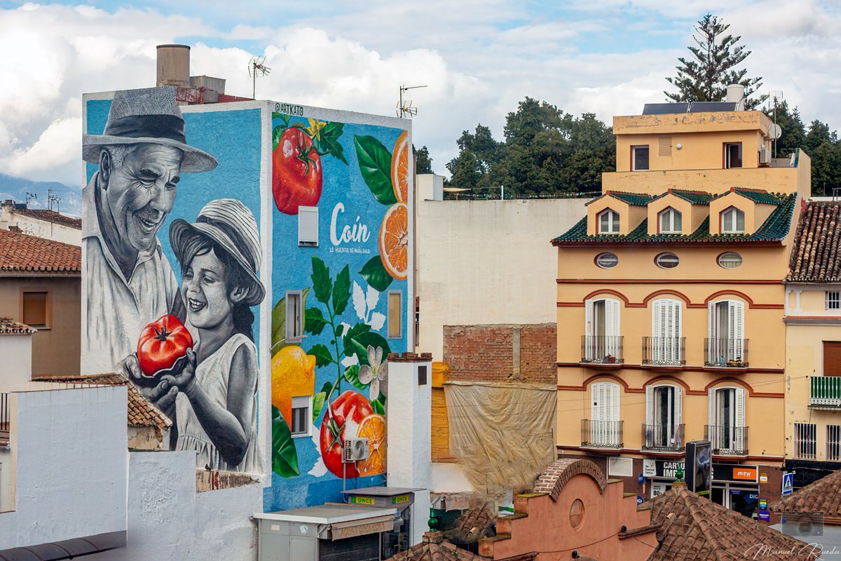 Obra del artista artkat0 donde refleja la imagen de un abuelo agricultor junto a su nieta recogiendo tomates del huerto en el lateral y en el frontal aparecen tomates y cítricos, como la naranja o el limón.
Coín
