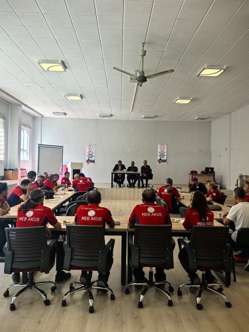 A large room with people wearing red "MEB AKUB" shirts sitting at tables, facing three individuals at a front table. Banners on the wall display "KONYA MER AKUB ARAMA KURTARMA BİRİMİ." Some people hold certificates, and Metin Tayarer is visible receiving a certificate with another man in a suit. The room has wooden flooring, ceiling lights, and a ceiling fan.