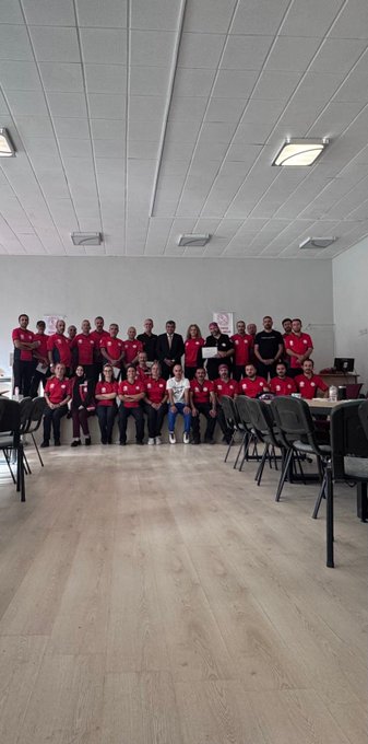 A large room with people wearing red "MEB AKUB" shirts sitting at tables, facing three individuals at a front table. Banners on the wall display "KONYA MER AKUB ARAMA KURTARMA BİRİMİ." Some people hold certificates, and Metin Tayarer is visible receiving a certificate with another man in a suit. The room has wooden flooring, ceiling lights, and a ceiling fan.